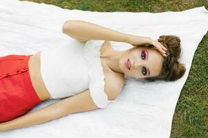 A beautiful young woman with bright makeup in the style of watermelon lies on a white blanket on the grass outdoors in summer photo