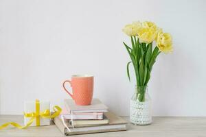 A mug, a lot of notepads in a stack, a bouquet of yellow spring tulips and a gift box on the desktop of a freelancer or blogger woman. Women's day, valentine's day or birthday. Front views photo