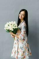 lado ver de un hermosa mujer con un ramo de flores de blanco tulipanes mirando a el flores y sonriente aislado en gris foto