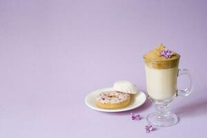 Beautiful dalgona drink a frothy coffee in a transparent mug and flowers of lilac on a purple background. Sweets on a plate. Donat and tender marshmallows. Copy space photo