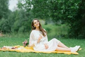 A happy smiling girl holds a glass of juice or wine and sits on a yellow blanket on a summer picnic outside the city photo