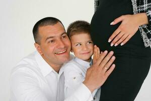 contento bebé chico, hijo y papá escucha y sostiene su manos en su embarazada de la madre estómago mientras esperando para un hermano o hermana foto