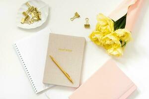 primavera femenino espacio de trabajo blogger cuadernos con un bolígrafo, un ramo de flores de amarillo tulipanes en un blanco antecedentes. de moda elegante plano laico foto