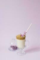 dalgona Coffee in a transparent Cup with a paper tube, a plate with lilac flowers on a lilac background with a copy space photo