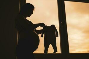 embarazada mujer en pie junto a ventana y participación bebé ropa. silueta de embarazada mujer. foto