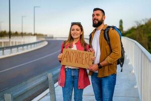 Happy couple hitchhiking on roadside trying to stop car. They are holding cardboard with inscription. photo