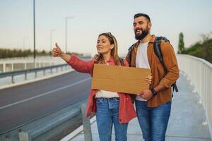 Happy couple hitchhiking on roadside trying to stop car. They are holding blank cardboard for your text. photo