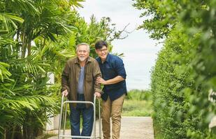 Caregiver is teaching old man to walk with walker, Senior asian father and middle aged son walk in the park, Elderly senior man with son walking outdoor photo