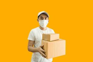 Young Asian delivery man wearing cap and white blank t-shirt holding parcel post box isolated on yellow background. express delivery service concept. photo