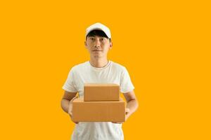 joven asiático entrega hombre vistiendo gorra y blanco blanco camiseta participación paquete o empaquetar enviar caja aislado en amarillo antecedentes. Rápido entrega Servicio concepto. foto