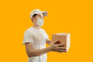 Young Asian delivery man wearing cap and white blank t-shirt holding parcel post box isolated on yellow background. express delivery service concept. photo