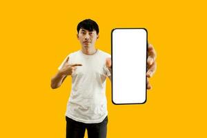 Asian Man Hand Pointing At Empty White Smartphone Screen on yellow Background. Cellphone Display Mock Up For Mobile App Advertisement. photo