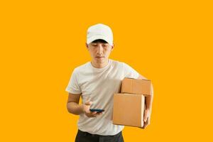 Young Asian delivery man wearing cap with white blank t-shirt holding smartphone and parcel post box isolated on yellow background. express delivery service concept. photo