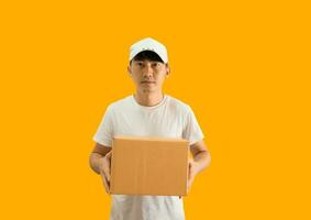 Young Asian delivery man wearing cap and white blank t-shirt holding parcel post box isolated on yellow background. express delivery service concept. photo