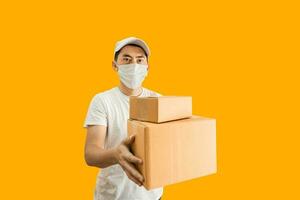 Young Asian delivery man wearing cap and white blank t-shirt holding parcel post box isolated on yellow background. express delivery service concept. photo