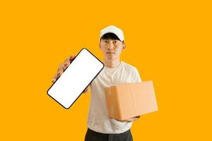 Young Asian delivery man wearing cap with white blank t-shirt holding smartphone and parcel post box isolated on yellow background. express delivery service concept. photo