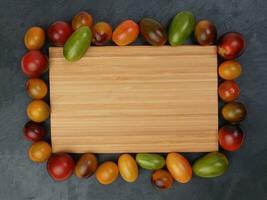 Red orange yellow green tomato mix variety on around wooden chopping board frame copy text space over black slate stone background photo
