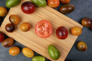 Red orange yellow green tomato mix variety on around wooden chopping board frame copy text space over black slate stone background photo