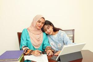 Two young Asian Malay Muslim woman wearing headscarf at home office student sitting at table talk mingle look at computer book document study discuss show affection care photo