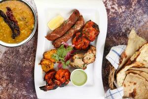 Mixed grill kebab chicken mutton beef prawn fish mint sauce lemon pickled onion bread basket chapati naan tandoori paratha dal lentil fry tadka copper pot on white dish over rustic background photo