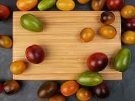 Red orange yellow green tomato mix variety on around wooden chopping board frame copy text space over black slate stone background photo