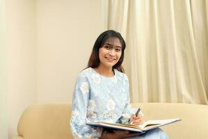 young Asian Malay Muslim woman wearing baju kurung dress at home sit rest on sofa read book notes write with pen look at camera photo