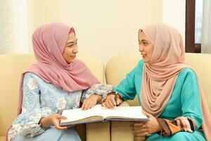 Two young Asian Malay Muslim woman wearing headscarf at home office student sitting on sofa talk mingle look at book document study discuss other smile happy photo