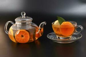 Liquid tea lemon orange slice green leaf cinnamon stick in transparent glass teacup saucer teapot kettle on black background photo