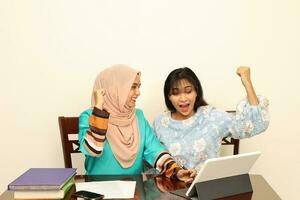 Two young Asian Malay Muslim woman wearing headscarf at home office student sitting at table talk mingle computer book document study discuss fist in air happy victory photo