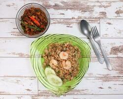Thai style stair fiend rice with prawn seafood cucumber slice garnish on green glass triangular plate basil minced chicken spicy red green chili fork spoon on rustic wood background flat lay top view photo