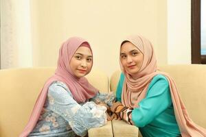 Two young Asian Malay Muslim woman wearing headscarf at home office student sitting on sofa talk mingle look at camera other smile happy photo