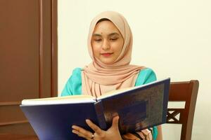 joven asiático malayo musulmán mujer vistiendo Pañuelo a hogar oficina estudiante sentado a mesa leer libro documento Nota estudiar sonrisa contento Mira foto