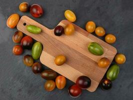 Red orange yellow green tomato mix variety on around wooden chopping board frame copy text space over black slate stone background photo
