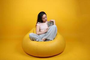 Beautiful young south east Asian woman sits on a yellow beanbag seat orange yellow color background pose fashion style elegant beauty mood expression rest relax read book think photo