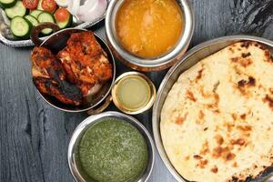 Spicy red Chicken grilled tikka tandoori nan bread green spinach curry yogurt sauce dal tomato cucumber onion salad set in metal stainless steel brass copper pot on black char wood background photo