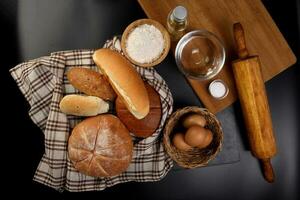 Freshly baked bread loaf bun roll round long mix verity wrapped in checkered kitchen fabric napkin towel wheat flower oil water salt eggs rolling pin slate stone over black background photo