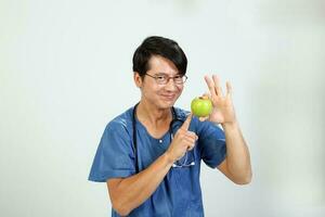 joven asiático masculino médico vistiendo delantal uniforme sayo estetoscopio participación señalando demostración comiendo sano verde manzana foto