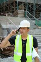 sur este asiático joven malayo hombre vistiendo blanco la seguridad casco amarillo chaleco hablando en teléfono foto