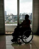 joven hombre sur este asiático malayo origen sentado en silla de ruedas en contra ventana ligero silueta. paralizado cintura abajo debido a accidente foto