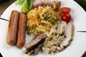 Mix sliced chicken beef steak sausage tomato onion garlic fry grill colorful pasta green salad cheese olive mint basil pesto sauce served on white plate over rustic wooden table photo