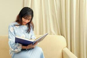 joven asiático malayo musulmán mujer vistiendo baju Kurung vestir a hogar sentar descanso en sofá leer libro notas escribir con bolígrafo pensar foto