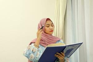 young Asian Malay Muslim woman wearing headscarf at home office next to window look at book document study think write photo