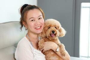 Beautiful attractive middle age southeast Asian woman sitting on sofa posing happy with small pet brown dog photo