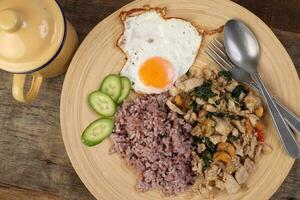 Thai fusion stair fried chicken with Thai basil and cashew nut with health brown rice fried egg sauce cucumber on woody plate over rustic wood background tin mug fork spoon photo