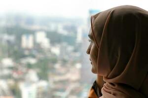 joven asiático malayo musulmán mujer vistiendo Pañuelo baju Kurung vestir a hogar en frente ventana Mira a paisaje urbano edificios al aire libre foto