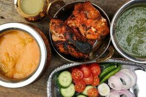 Spicy red Chicken grilled tikka tandoori nan bread green spinach curry yogurt sauce dal tomato cucumber onion salad set in metal stainless steel brass copper pot on rustic vintage wood background photo