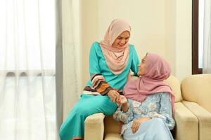 Two young Asian Malay Muslim woman wearing headscarf at home office student sitting on sofa talk mingle smile happy photo