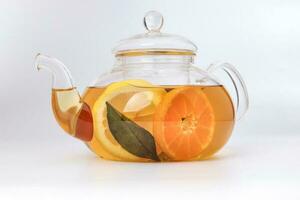 Liquid tea lemon orange slice green leaf cinnamon stick in transparent glass teapot kettle on white background photo