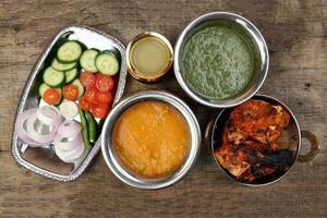 Spicy red Chicken grilled tikka tandoori nan bread green spinach curry yogurt sauce dal tomato cucumber onion salad set in metal stainless steel brass copper pot on rustic vintage wood background photo