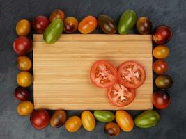 Red orange yellow green tomato mix variety on around wooden chopping board frame copy text space over black slate stone background photo